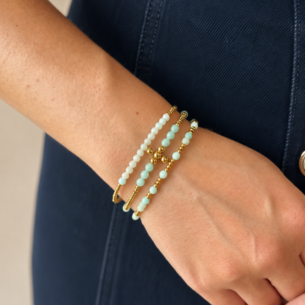 A close-up of a person's wrist wearing three beaded bracelets: two light aqua beads with gold spacer beads and one white bead bracelet with gold accents, worn over a dark navy top.