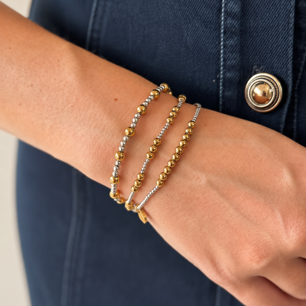 Three beaded bracelets in gold and silver tones worn stacked on a person's wrist, shown against a dark denim jacket.