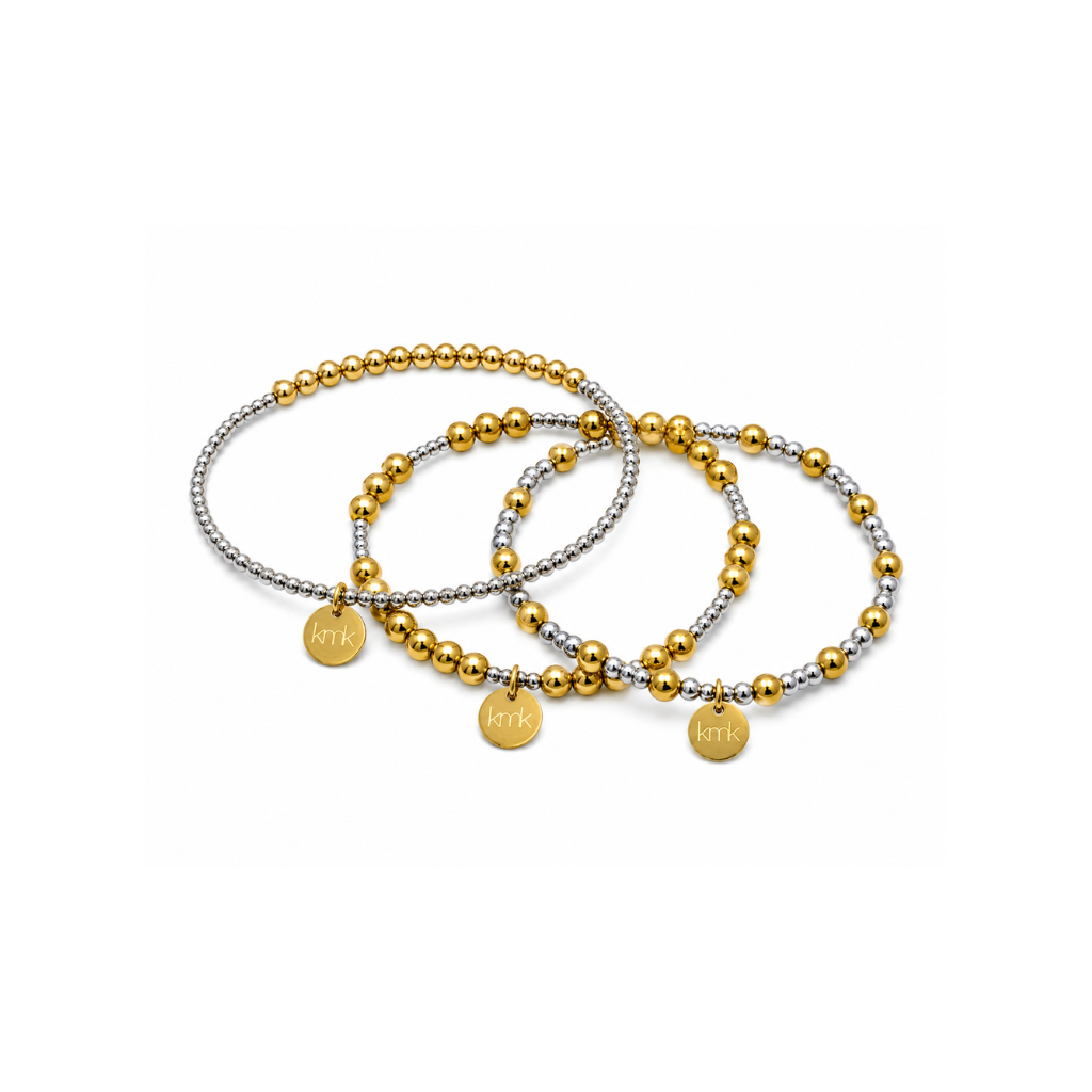 Three beaded stretch bracelets with gold and silver metallic beads and small round gold charms engraved with 'KMK', arranged on a plain white background.