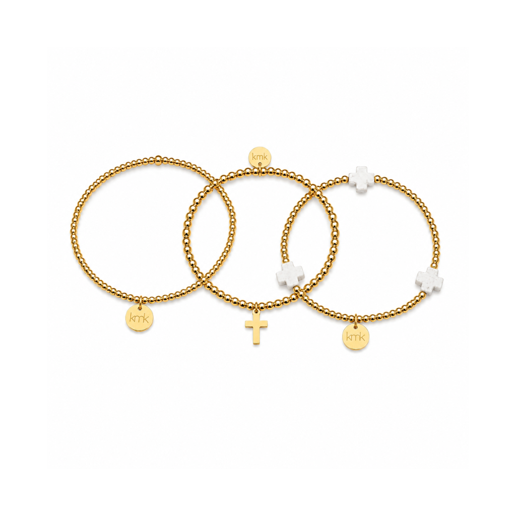 Three delicate gold-toned beaded bracelets with small charms, including a cross, displayed on a clean white background.