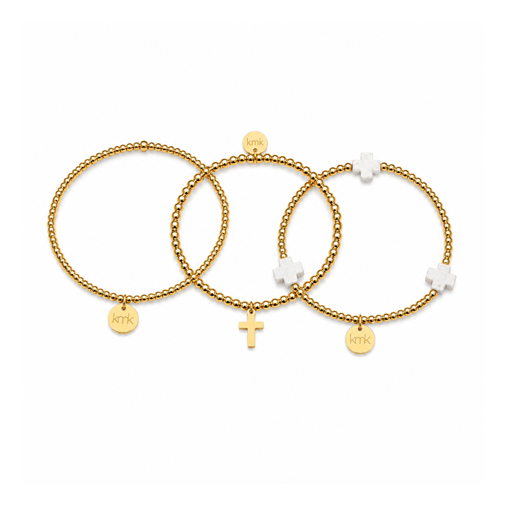 Three gold beaded stretch bracelets with small charms, including a cross, a round engraved disc, and white flower-shaped beads, displayed on a clean white background.