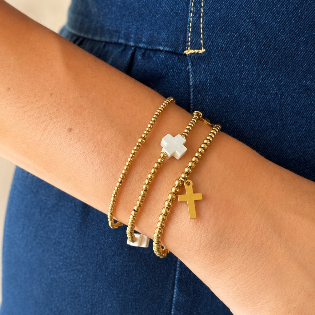 A close-up of a person's wrist wearing three delicate gold bracelets, including two beaded chains and two with cross charms (one gold, one white), against a blue denim background.