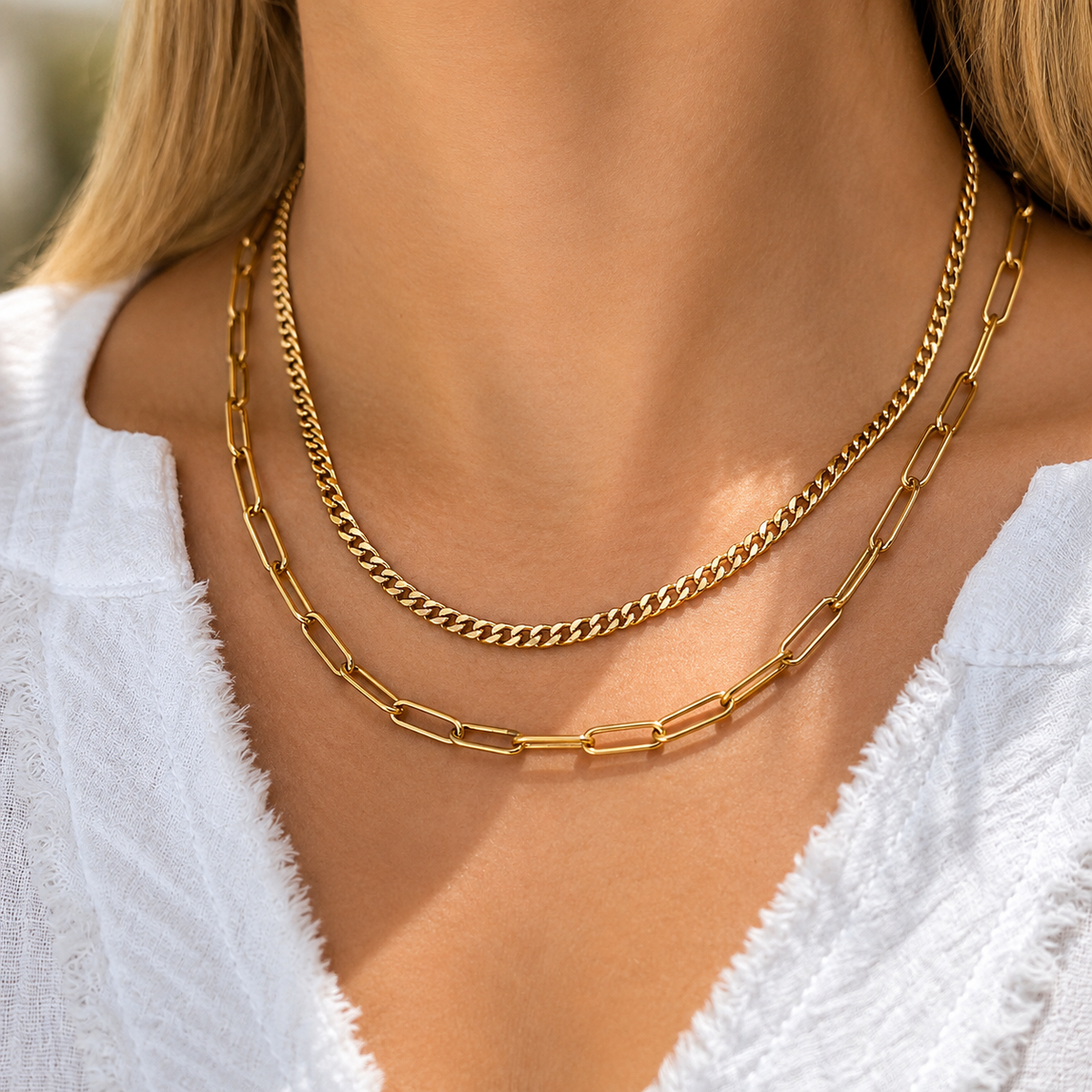 A close-up of a model's neck and chest wearing two layered gold necklaces: a fine curb chain and a thicker paperclip-style chain, paired with a white textured top.