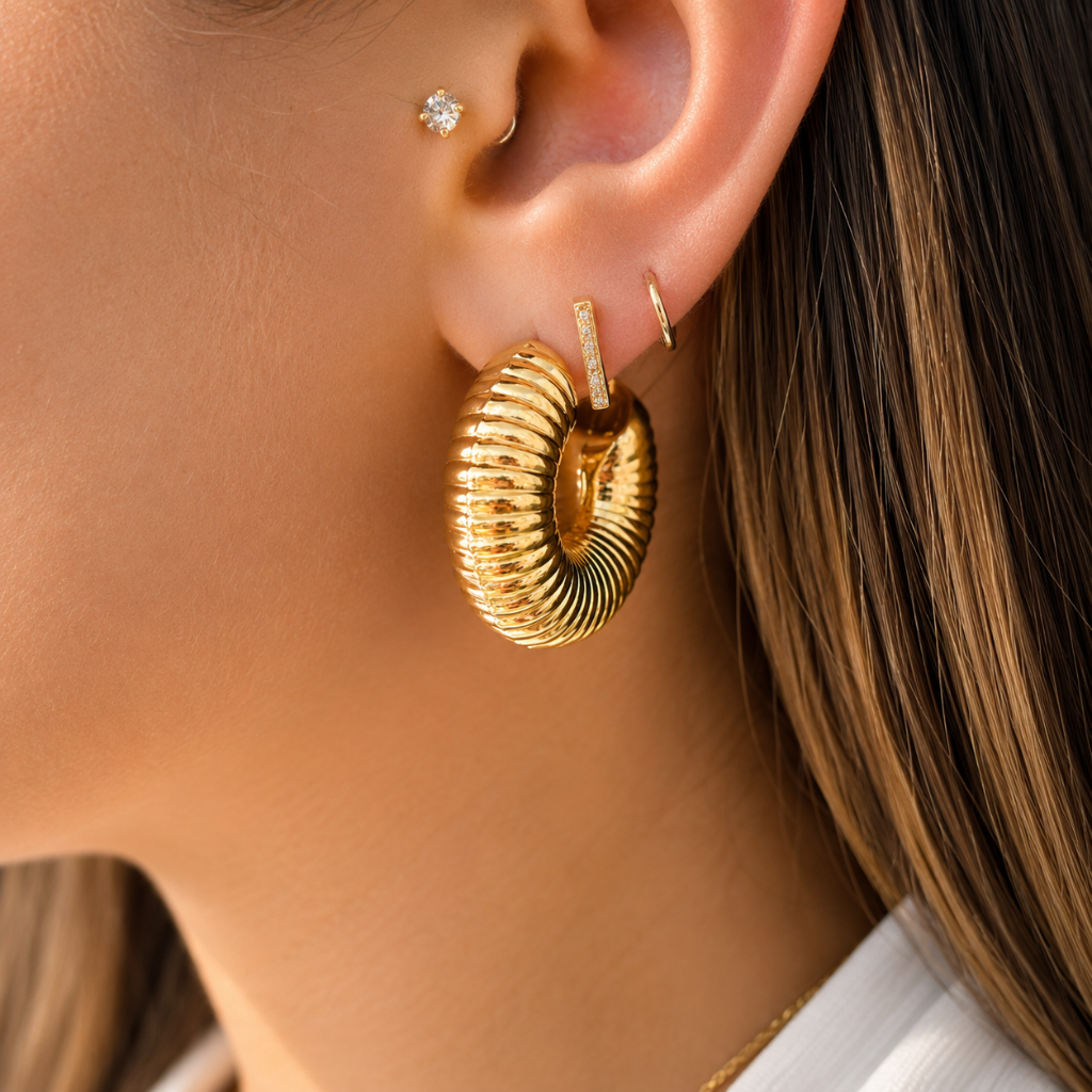 A close-up of a model's ear showcasing a large, textured gold hoop earring, along with smaller gold ear piercings.