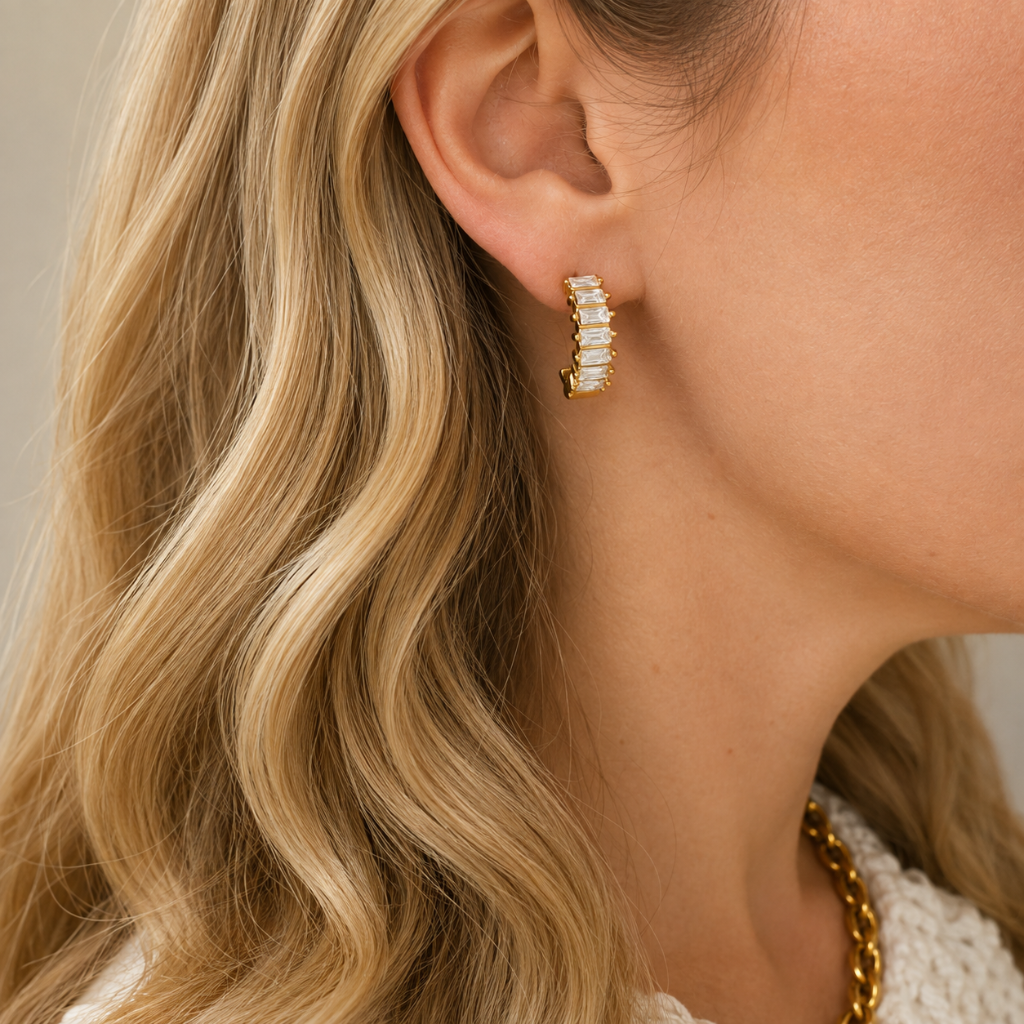A close-up of a model's ear wearing a gold crescent-shaped hoop earring with baguette-cut clear crystals, styled with a gold chain necklace and blonde wavy hair.