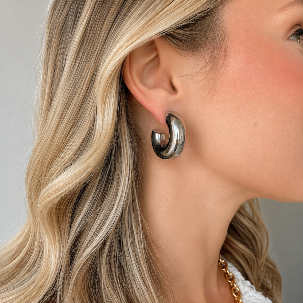 A close-up of a woman's ear wearing a polished silver crescent-shaped hoop earring, with her blonde hair and neck visible against a soft gray background.
