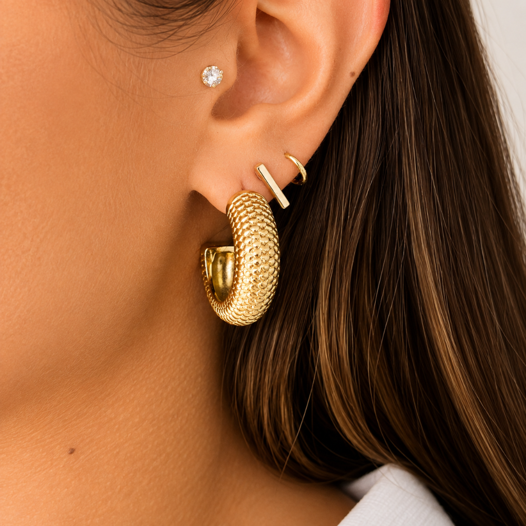 A close-up of a person's ear showcasing a gold textured hoop earring, a small gold bar stud, and a tiny gold huggie hoop, alongside a diamond stud earring.