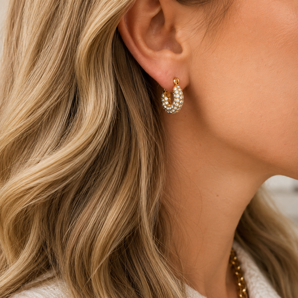 A close-up of a woman's ear wearing a small gold crescent-shaped hoop earring adorned with white pearls, set against her blonde hair and neck.