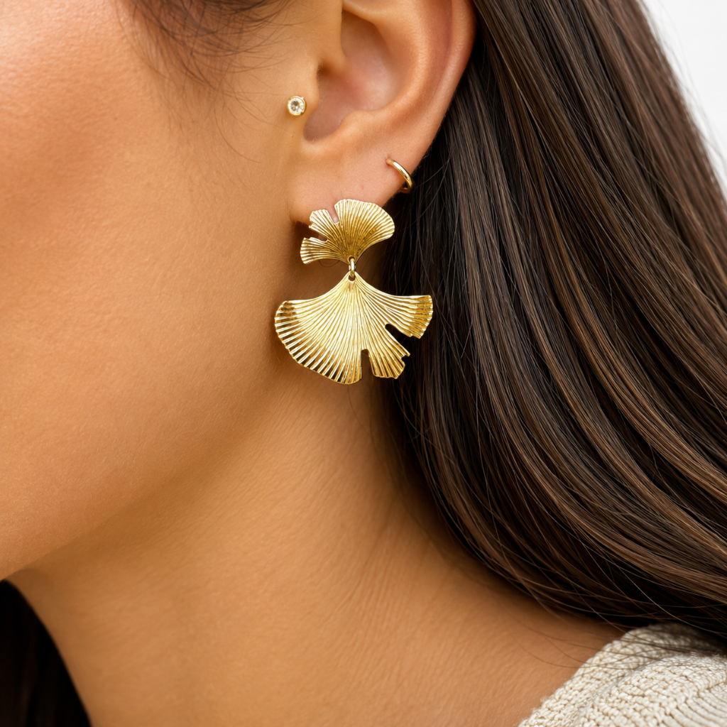 A close-up of a model's ear wearing a large, textured gold ginkgo leaf drop earring, along with smaller gold hoop earrings.