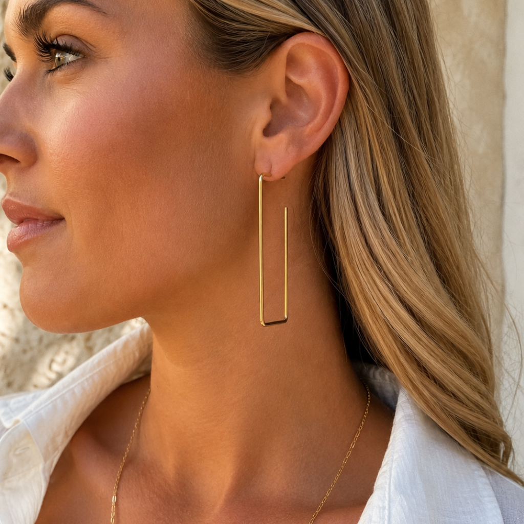 A close-up side profile of a woman wearing a delicate gold geometric hoop earring and a thin gold chain necklace, dressed in a white collared shirt.