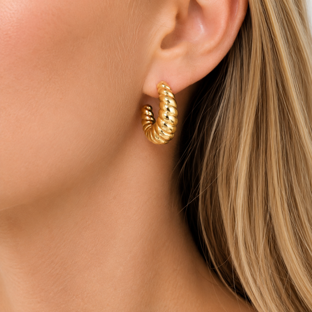 A close-up of a model's ear wearing a small twisted gold huggie hoop earring.