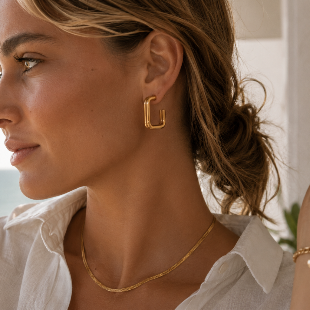 A close-up lifestyle shot of a woman wearing a gold geometric U-shaped hoop earring and a delicate gold chain necklace, paired with a white collared shirt.