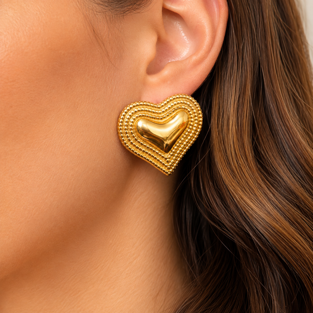 A close-up of a woman's ear wearing a textured gold heart-shaped stud earring with concentric beaded borders and a polished central heart.
