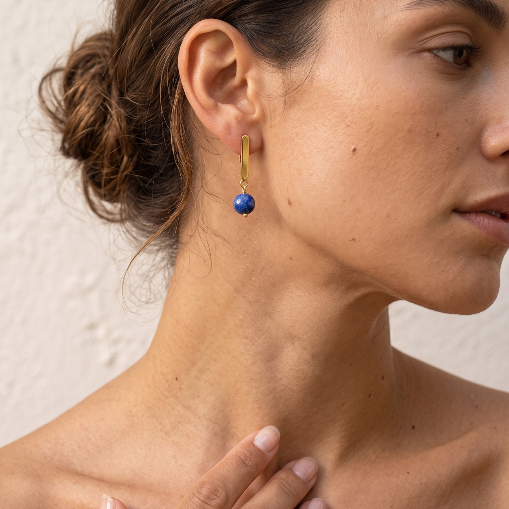 A close-up of a woman wearing a gold dangle earring with a small round blue bead, shown from the side against a light textured wall.