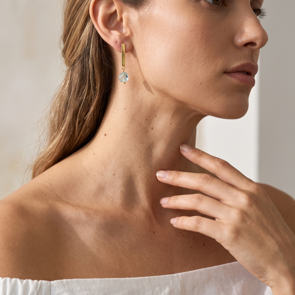 A close-up lifestyle shot of a woman wearing a delicate gold dangle earring with a faceted light blue crystal drop, set against a soft, warm neutral background.