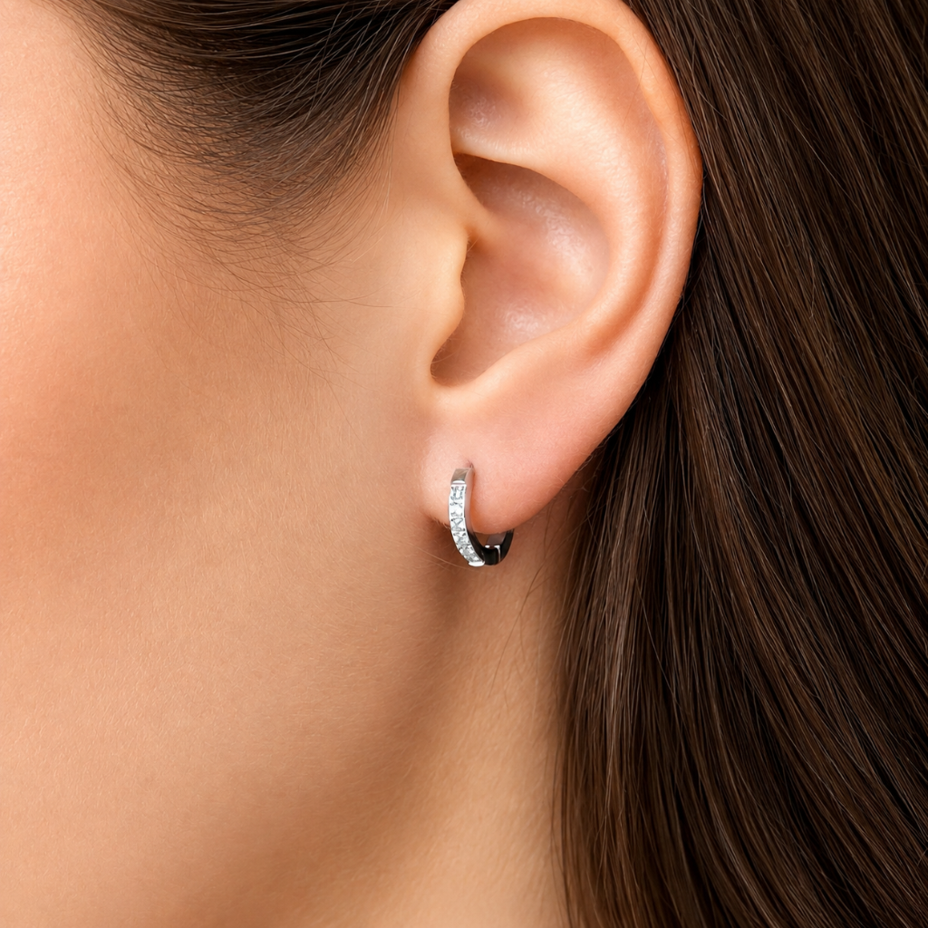 A close-up of a model's ear wearing a small silver huggie hoop earring set with clear stones, against her dark brown hair and skin.