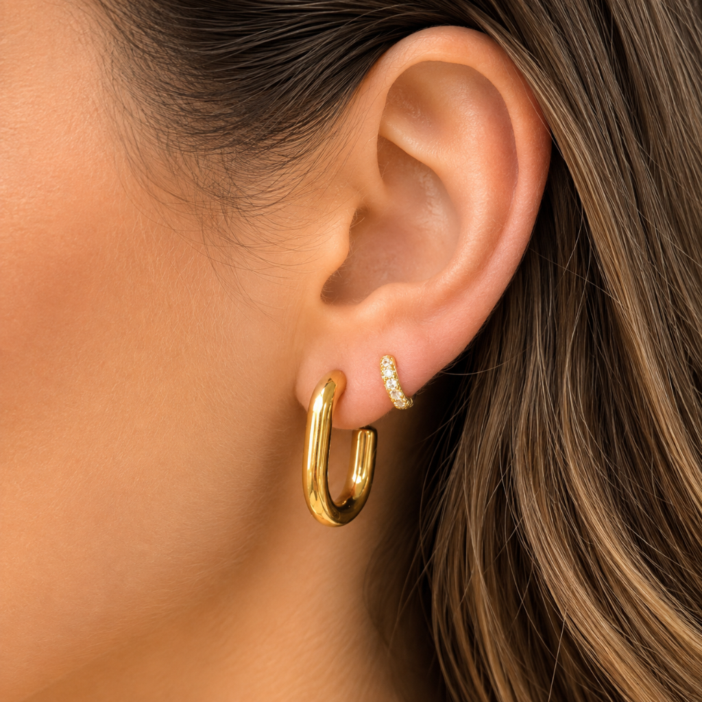 A close-up of a model's ear wearing a thick gold hoop earring and a delicate gold stud with small clear stones.