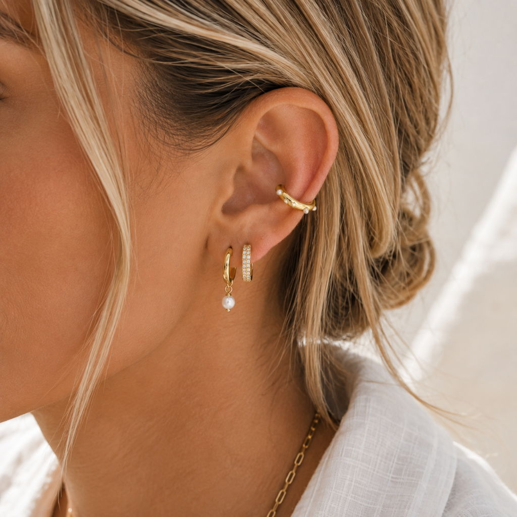 A close-up of a woman's ear wearing a stack of gold earrings, including a small gold hoop with a white pearl drop and a gold huggie hoop with tiny clear stones, against a bright neutral background.