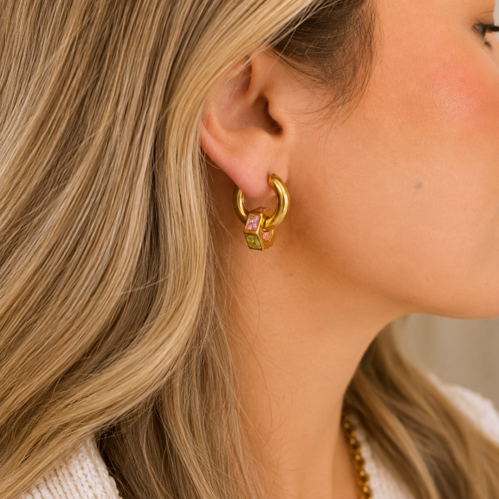 A close-up side profile of a woman with blonde hair wearing a gold huggie hoop earring adorned with small pink and green gemstone cubes.