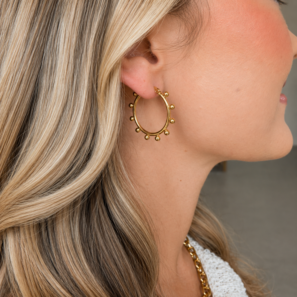 A close-up side profile of a woman wearing a gold hoop earring adorned with small dangling beads, styled with blonde hair and a white textured top.