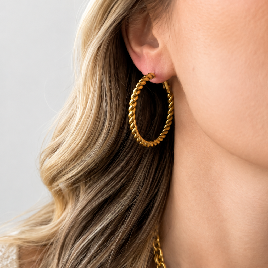A close-up of a woman's ear wearing a thick, twisted gold hoop earring, with her blonde hair and neck visible against a soft light gray background.