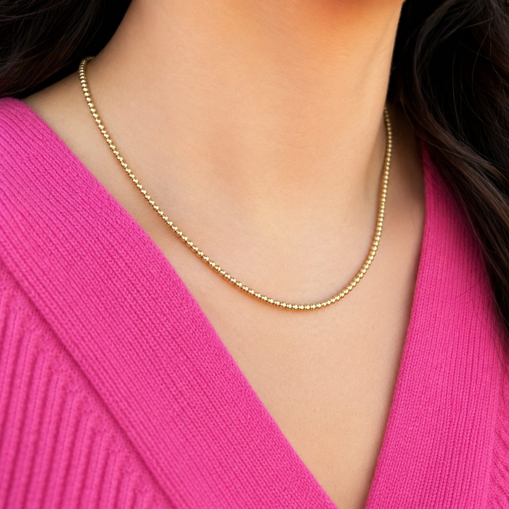 A close-up of a delicate gold beaded chain necklace worn by a model in a bright pink ribbed sweater.