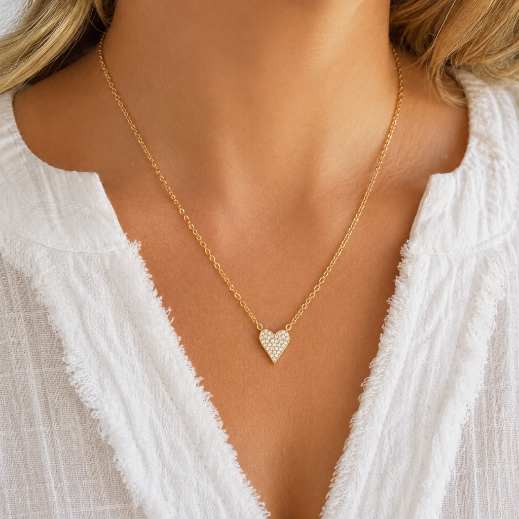 A close-up of a model wearing a delicate gold chain necklace with a small heart-shaped pendant adorned with tiny clear stones, paired with a white textured top.