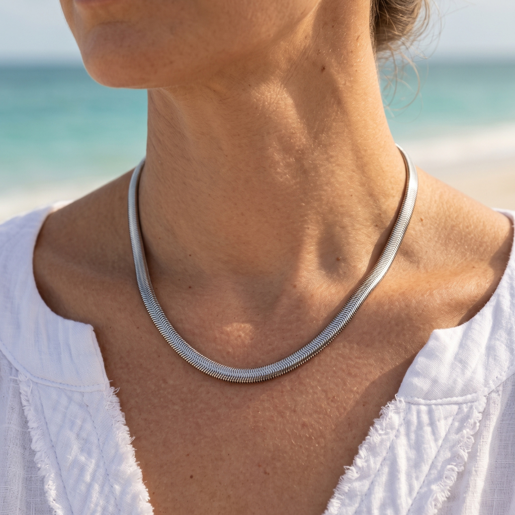 A close-up of a woman's neck and upper chest wearing a thin silver snake chain necklace, with a blurred beach and ocean background.
