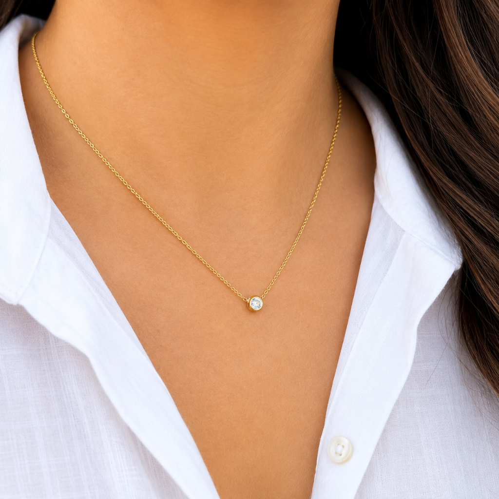 A close-up of a model wearing a delicate gold chain necklace with a small bezel-set clear gemstone pendant, styled with a white button-down shirt.