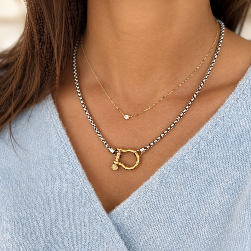 A close-up of a person wearing layered necklaces: a silver chain necklace with a gold D-shaped pendant and a delicate gold chain with a small round stone, over a light blue V-neck top.