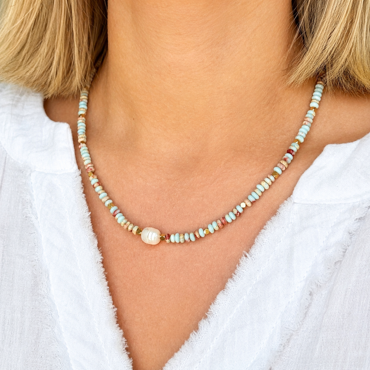 A close-up of a woman wearing a delicate beaded necklace with a central white pearl. The necklace features small, multi-colored beads in pastel shades of blue, green, pink, and orange.