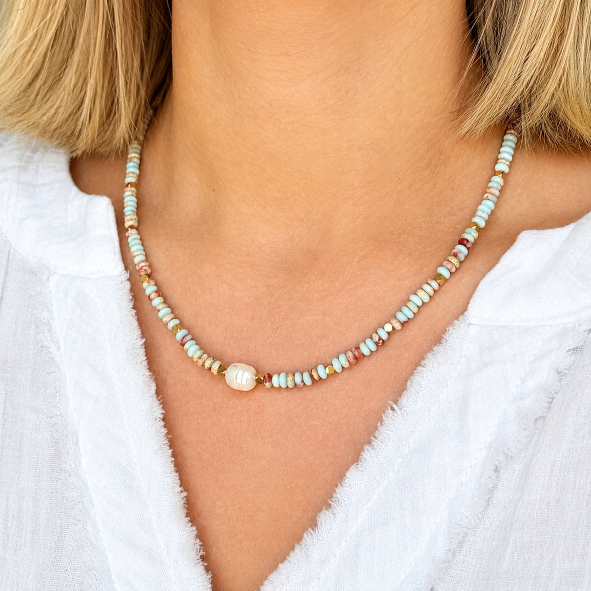A close-up of a woman wearing a delicate beaded necklace with a central white pearl. The necklace features small, multi-colored beads in pastel shades of blue, green, pink, and orange.