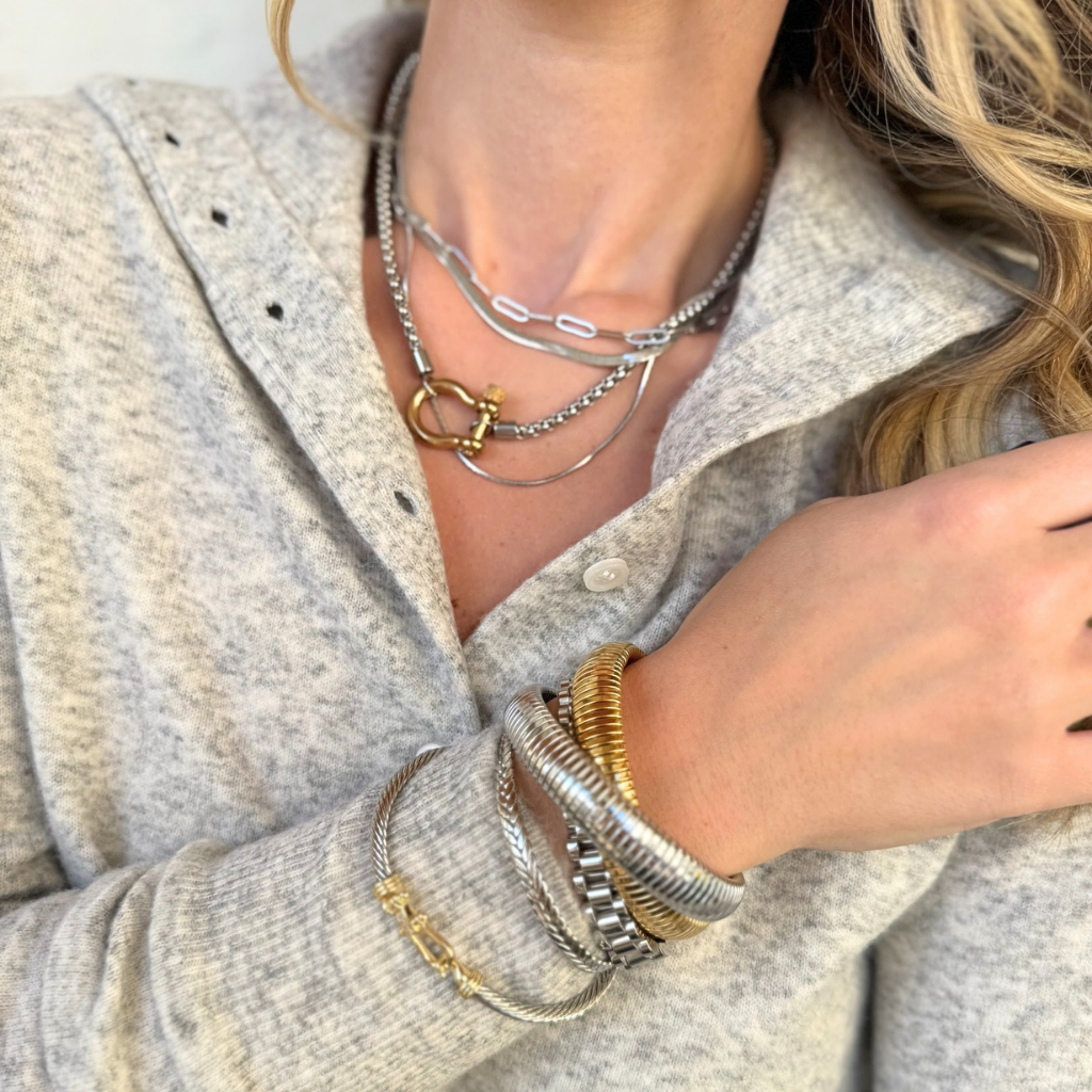 A close-up of a person wearing layered silver and gold necklaces with stacked bracelets on the wrist, paired with a light gray knit button-up shirt.