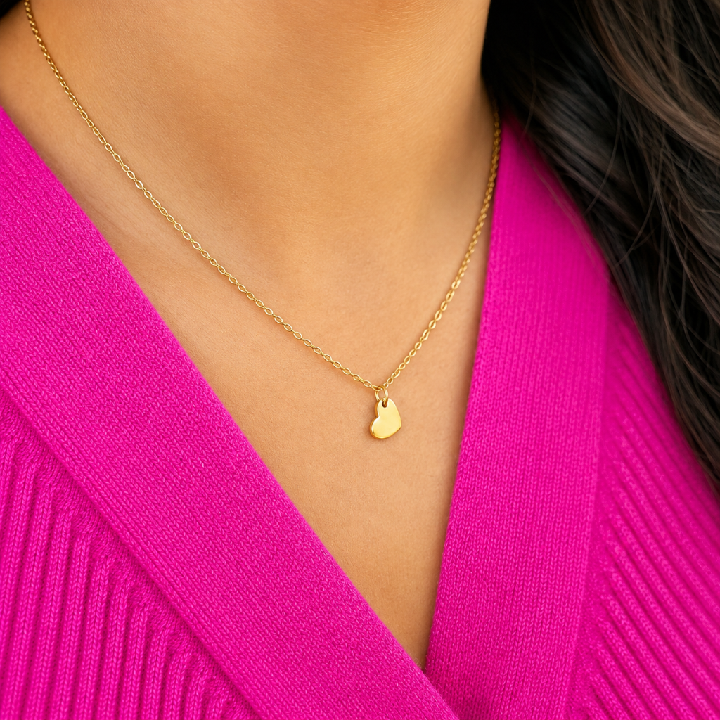 A close-up of a person wearing a delicate gold chain necklace with a small heart-shaped pendant, styled with a bright pink ribbed knit top.