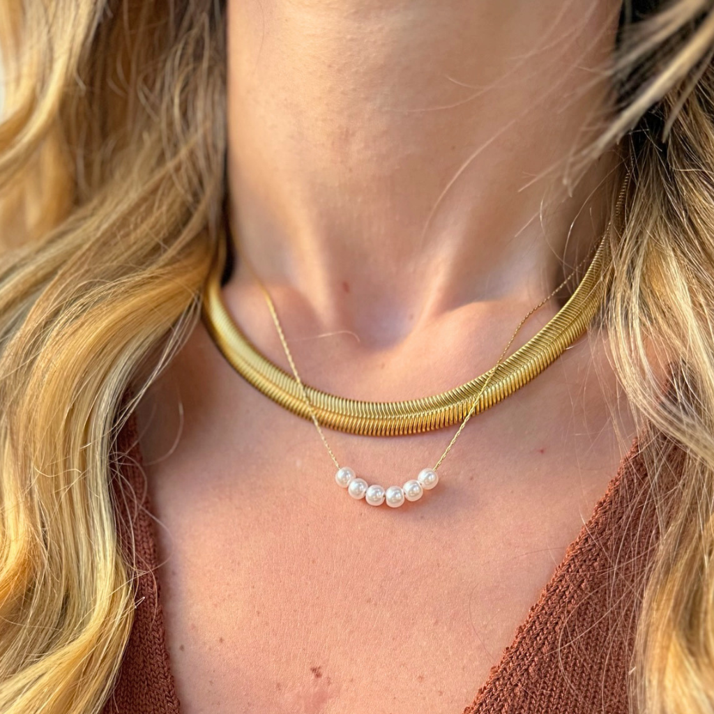 A close-up of a woman's neck and chest wearing a layered necklace set: a wide gold snake-style choker and a delicate gold chain with a row of small white pearls.