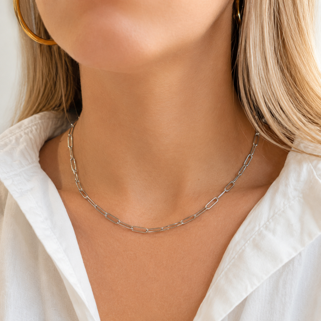 A close-up of a model wearing a silver paperclip chain necklace, styled with a white collared shirt and gold hoop earrings.