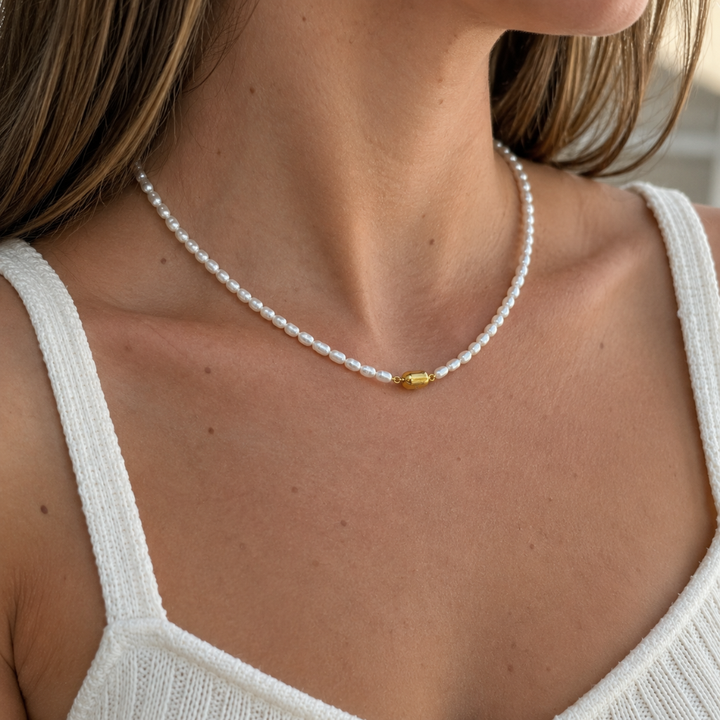 A close-up of a woman's neck and chest wearing a delicate white pearl necklace with a small gold accent bead at the center, paired with a white ribbed tank top.
