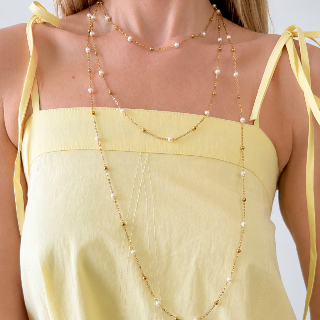 A close-up of a model wearing three layered gold chain necklaces with small white pearls, styled with a pale yellow tie-strap top.