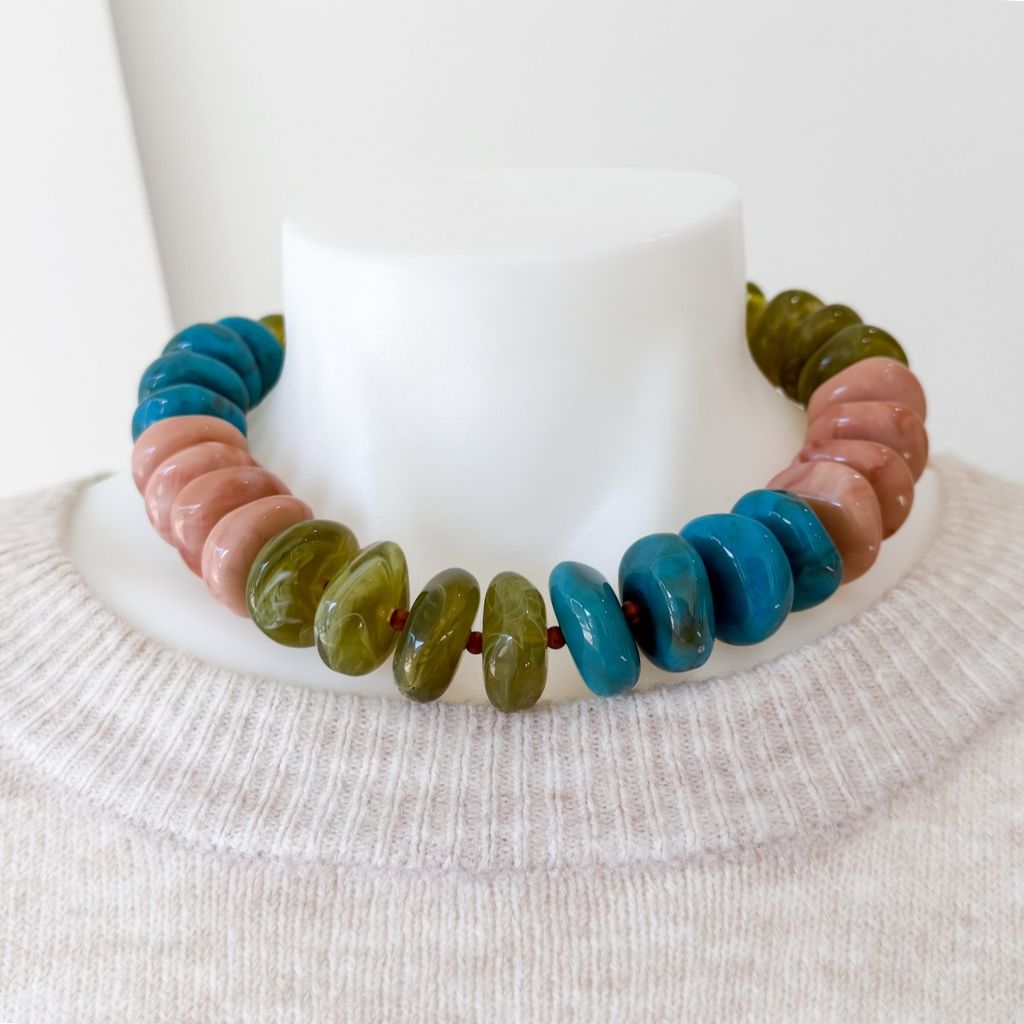 A beaded necklace made of alternating turquoise blue and olive green polished stone beads, displayed on a white mannequin bust wearing a cream knit sweater.