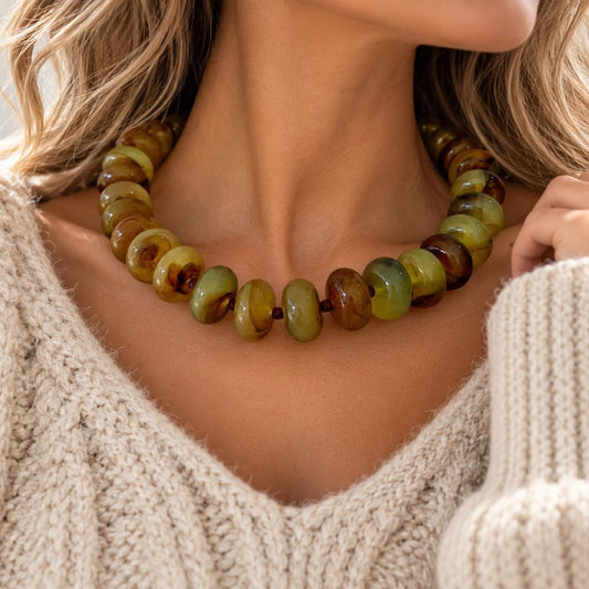 A close-up of a woman wearing a chunky beaded necklace made of olive green and brown polished stone beads, paired with a cream-colored knit sweater.