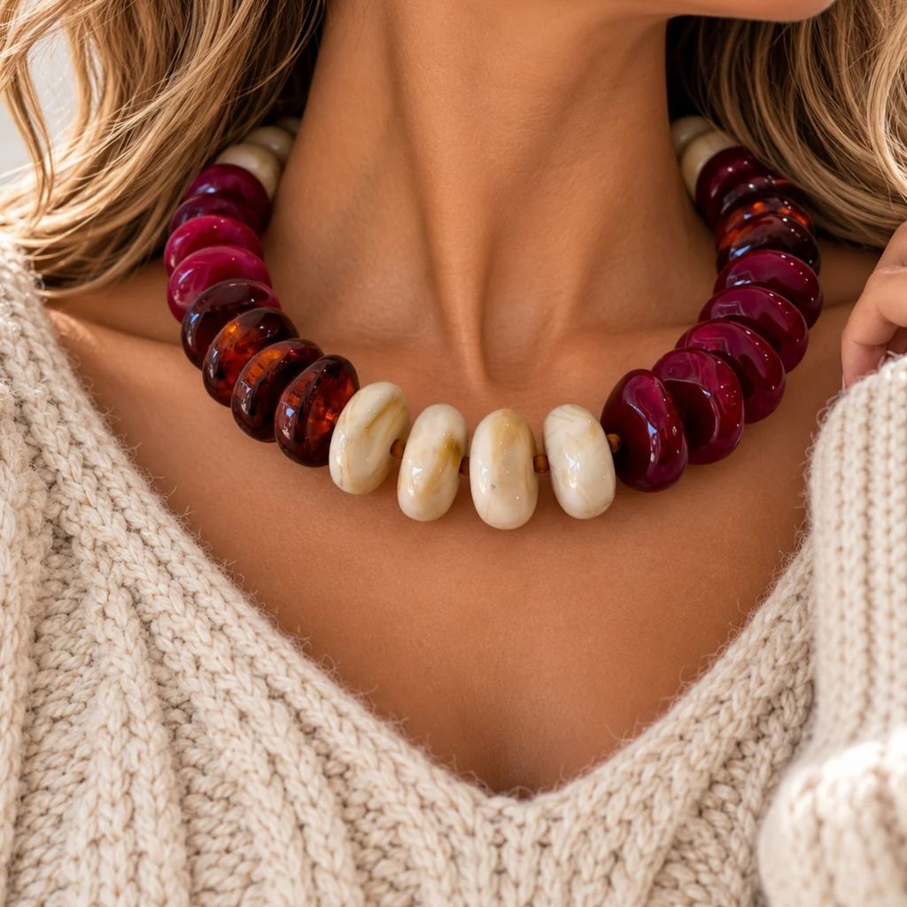 A close-up of a woman wearing a chunky statement necklace made of glossy dark red and amber-brown disc beads with five large cream-colored oval beads at the front, paired with a cream cable-knit sweater.