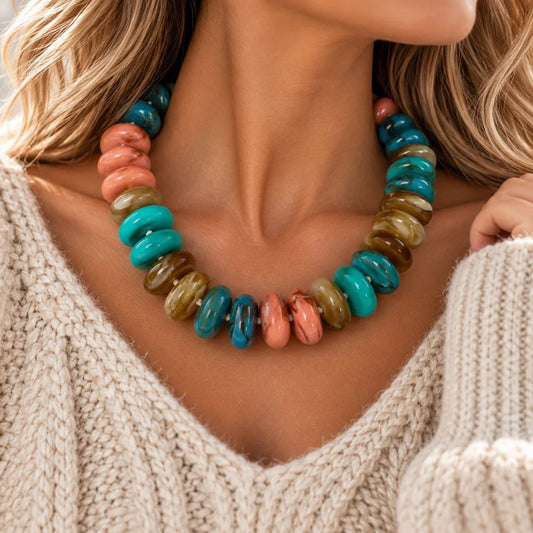 A close-up of a woman wearing a chunky beaded necklace made of glossy, disc-shaped beads in turquoise, coral pink, olive green, and brown, paired with a cream knit sweater.