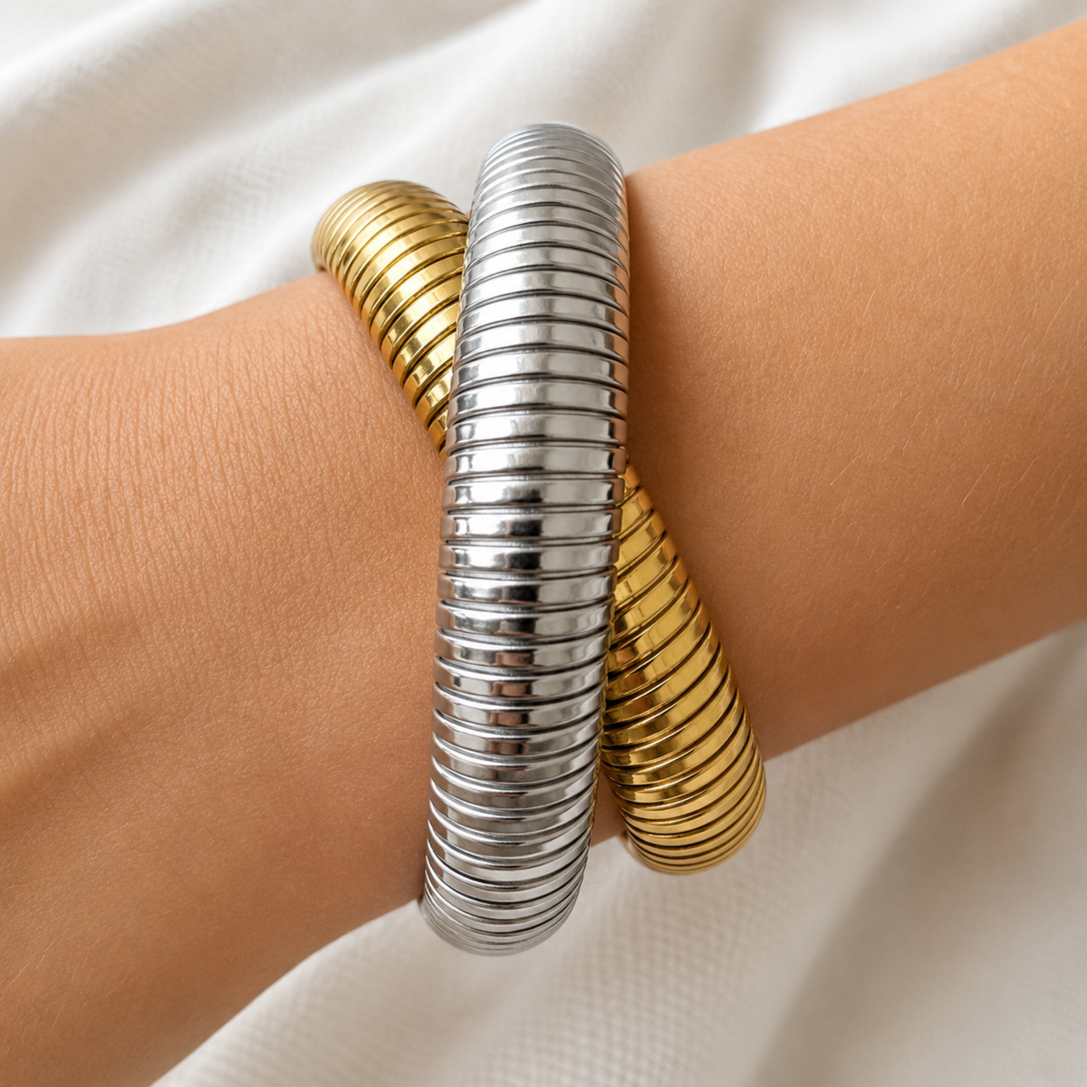 Two metallic bangles, one silver and one gold, wrapped together on a person's wrist against a soft white fabric background.