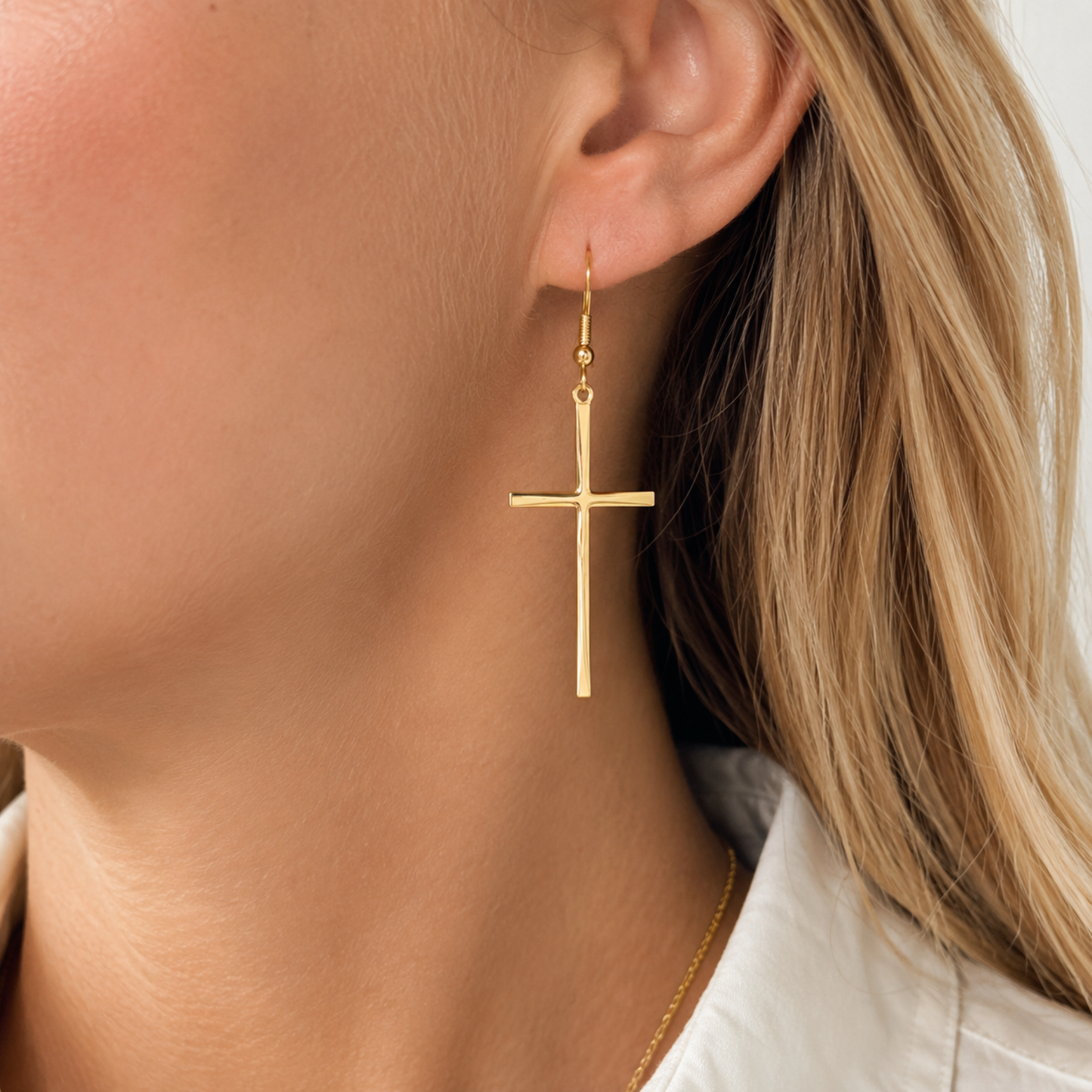 A close-up of a woman's ear and neck wearing a delicate gold cross dangle earring, with her blonde hair and white shirt visible.