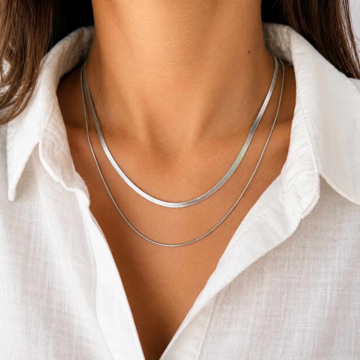 A close-up of a person's neck and collarbone area wearing two layered silver-toned chain necklaces over a white collared shirt.